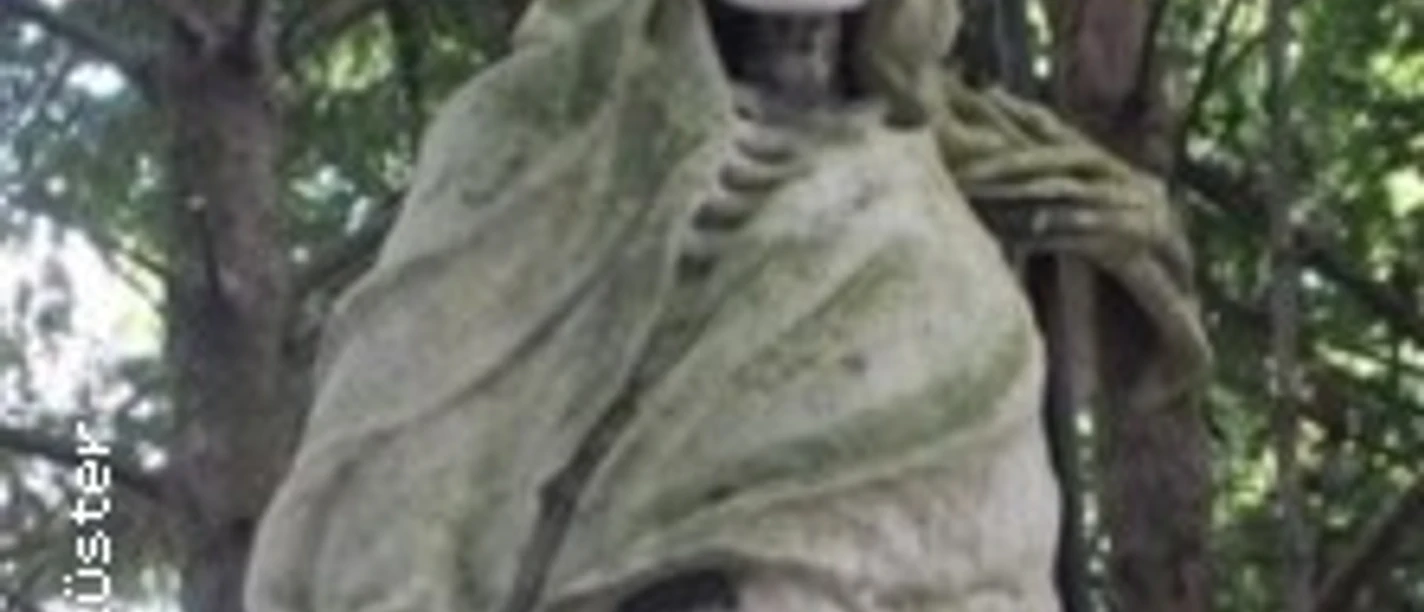 Verwitterte Skelettfigur aus Stein in Gewand, umgeben von dichtem Grün