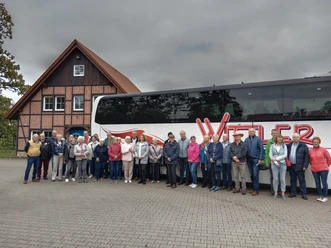 Vor einem modernen Reisebus der Firma 'Witter' posiert eine gemischte Gruppe von etwa 24 Personen, darunter Männer und Frauen unterschiedlichen Alters. Der Hintergrund zeigt ein traditionelles Fachwerkhaus, während der Himmel grau und wolkenverhangen wirkt.