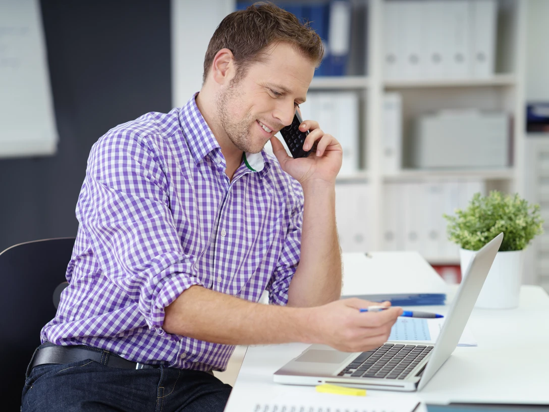 Dienstleistungen Ein Mann in kariertem Hemd telefoniert lächelnd am Schreibtisch vor einem Laptop.