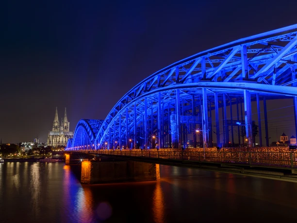 Weltdiabetestag Kölner Dom und Hohenzollernbrücke bei Nacht, beleuchtet in Blau, spiegeln sich im Rhein.