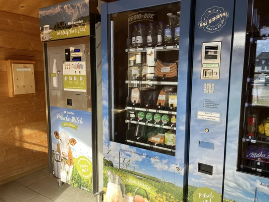 Milchautomat Bichlerhof, Farchant im kleinen Holzhäusl steht linkerhand ein Milchautomat mit Flaschen, rechts davon zwei Automaten mit Yogurt, Wurst und Fleischauswahl, vakumiert. in der Mitte findet sich der Bezahlautomat