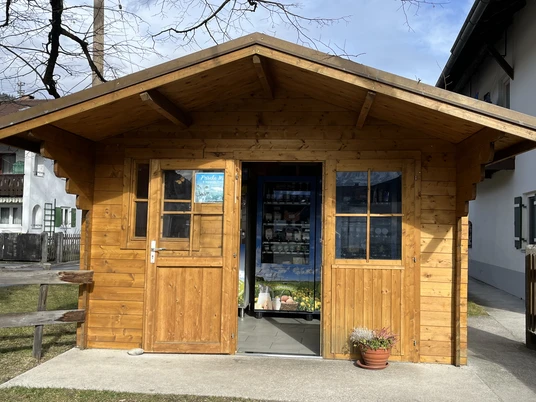 Milchautomat Bichlerhof, Farchant Aussenansicht des 24-Stundenautomats. Aus Holz gestaltete kleine Hütte mit 2 Fenstern, 1 Türe. Davor steht eine Schale mit Blumen. Im Inneren des Häuschens erkennt man die Automaten
