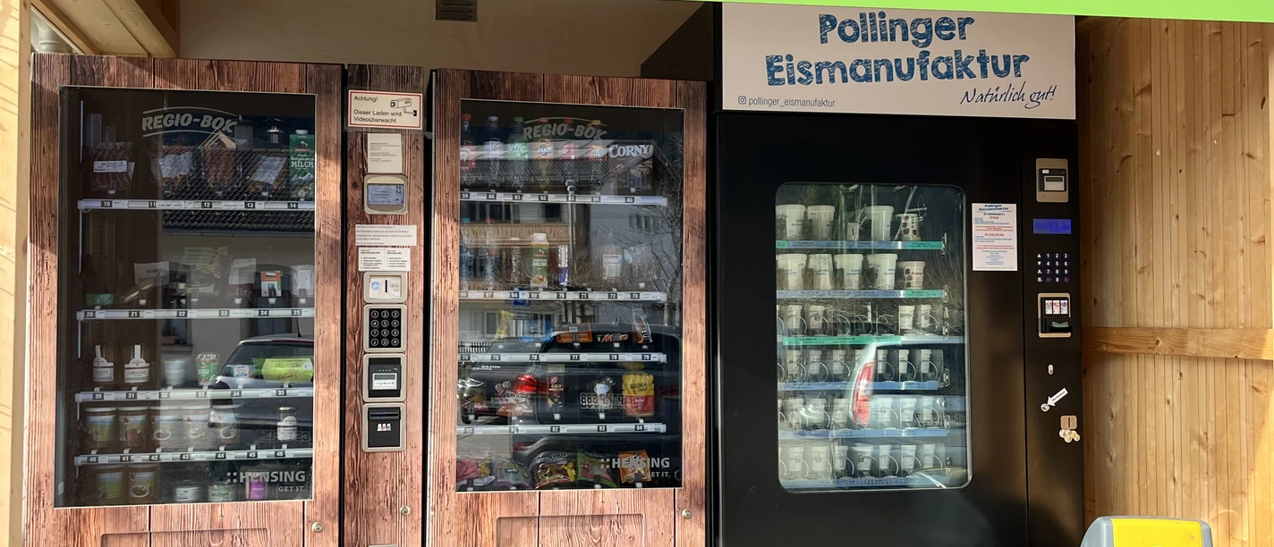 Lebensmittelautomat in FArchant 2-türiger Kühlschrank mit verschiedenen regionalen Lebensmitteln sowie ein Maschine für Softeis, darüber steht auf grünem Banner : 24 Stunden einkaufen zu bezahlen in bar und mit Karte