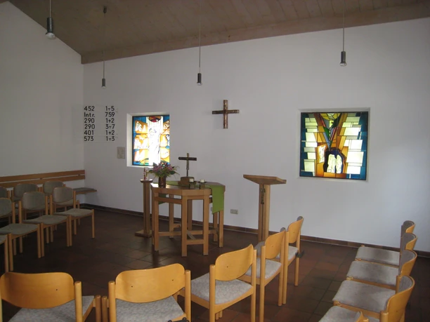 Innenansicht mit Holzstühlen und- Altar, 2 Fenster mit Einlegearbeiten und Holzkreuz an der Wand