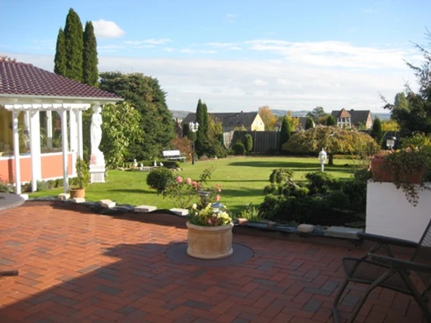 Gepflegter Garten mit Rasenfläche, umgeben von Bäumen, Blumen und einer Terrasse bei sonnigem Wetter.