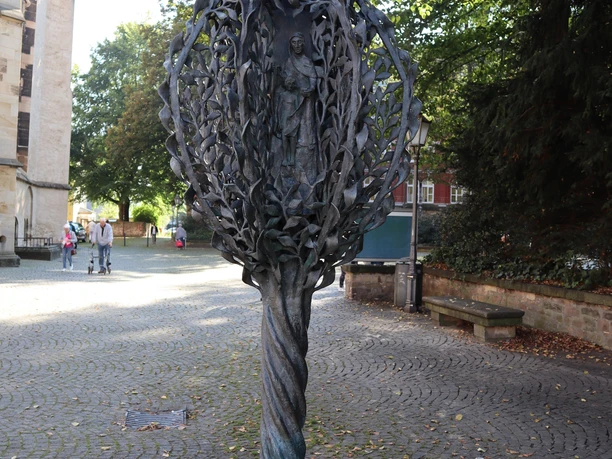 Abtei-Stele (1).JPG Stele mit filigranen Pflanzenmustern und Figuren, von Bäumen umgeben auf einem gepflasterten Platz.