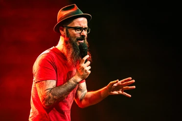 Jens Kotalla trägt einen Hut und roten T-Shirt, während er mit Mikrofon spricht, intensiv und fokussiert.