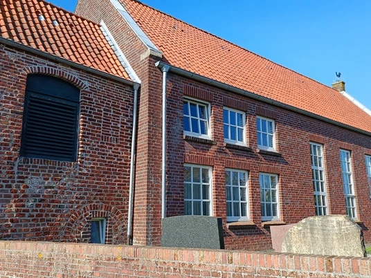 Ev.-ref. Kirche Hamswehrum Backsteinkirche mit roten Dachziegeln und Sprossenfenstern, umgeben von Grabsteinen unter blauem Himmel.