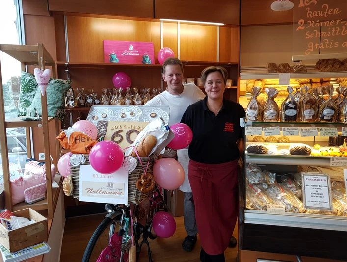 Café & Konditorei Nollmann Team Eine Bäckerei feiert 60 Jahre. Zwei Personen lächeln neben einem dekorierten Fahrrad voller Brot.