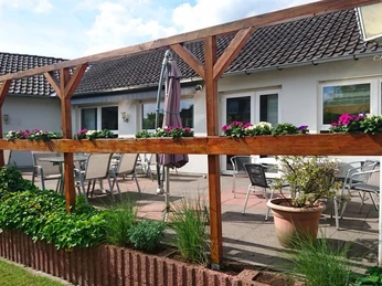 Café & Konditorei Nollmann Terrasse Terrasse mit Holzelementen, Blumenkästen und Sitzgelegenheiten vor einem weißen Gebäude mit Dachziegeln.