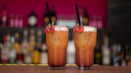 Zwei bunte Cocktails mit Kirschen als Dekoration stehen auf einem Tresen vor einer unscharfen Bar.