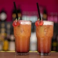Zwei bunte Cocktails mit Kirschen als Dekoration stehen auf einem Tresen vor einer unscharfen Bar.