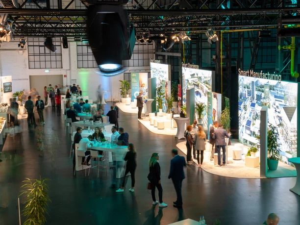 Fujitsu Smart Solutions Various groups of people are gathered in a large, modern event room. Bright lights and large screens presenting various topics related to smart manufacturing dominate the scene. The atmosphere is lively, with conversations and networking at bar tables.