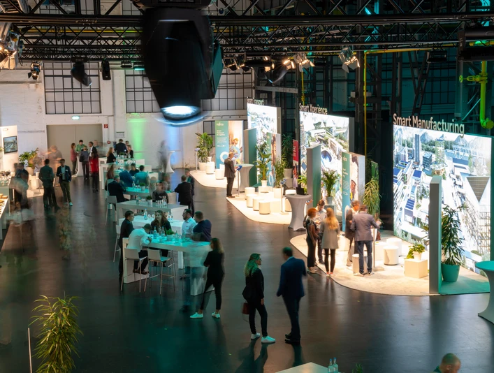 Fujitsu Smart Solutions In einem großen, modernen Veranstaltungsraum sind verschiedene Gruppen von Menschen versammelt. Helle Lichter und große Bildschirme, die verschiedene Themen rund um "Smart Manufacturing" präsentieren, dominieren die Szenerie. Die Atmosphäre ist belebt, mit Gesprächen und Networking an Stehtischen.Various groups of people are gathered in a large, modern event room. Bright lights and large screens presenting various topics related to smart manufacturing dominate the scene. The atmosphere is lively, with conversations and networking at bar tables.