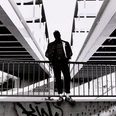 PoebelMC_Pressefotos2021_FotocreditMarshlCeronPalomino_5-scaled.jpg Ein Mann in urbaner Kulisse steht vor Geländer unter Brückenstruktur, kontrastreich in Schwarzweiß.A man in an urban setting stands in front of railings under a bridge structure, rich in contrast in black and white.Een man in een stedelijke omgeving staat voor leuningen onder een brugstructuur, contrastrijk in zwart-wit.