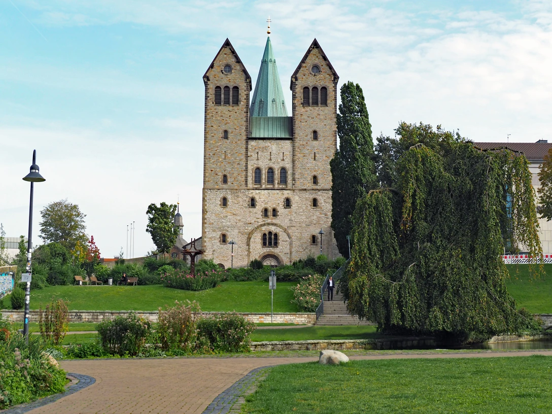 Abdinghofkirche in Paderborn vor blauem Himmel, umgeben von grünen Rasenflächen und Bäumen.