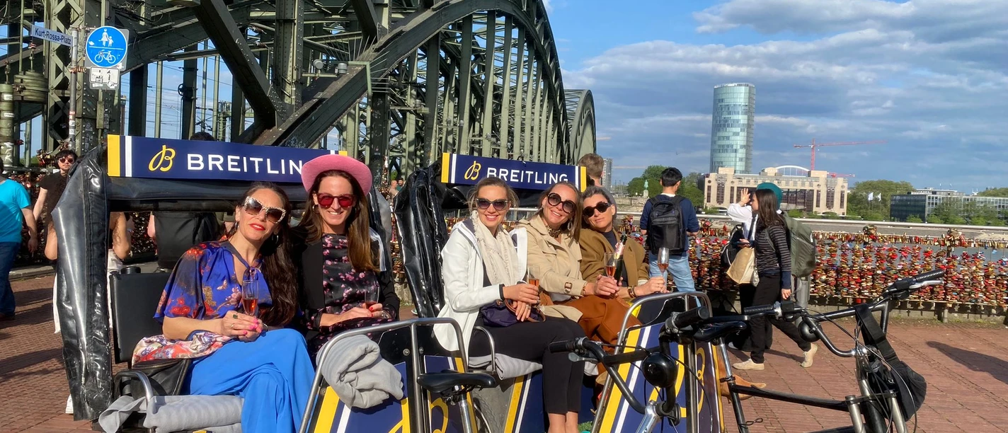 Rikolonia Champagnerfahrt.jpg Fünf Frauen sitzen in einer Fahrrad-Rikscha vor einer Brücke mit Vorhängeschlössern und genießen den Ausblick.</p>