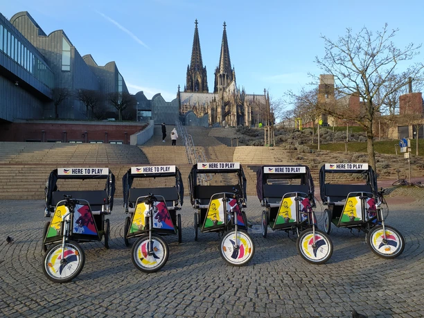 Rikolonia_Event Branding.jpg Die farbenfrohen Fahrradrikschas stehen vor dem eindrucksvollen Kölner Dom in der Abenddämmerung.