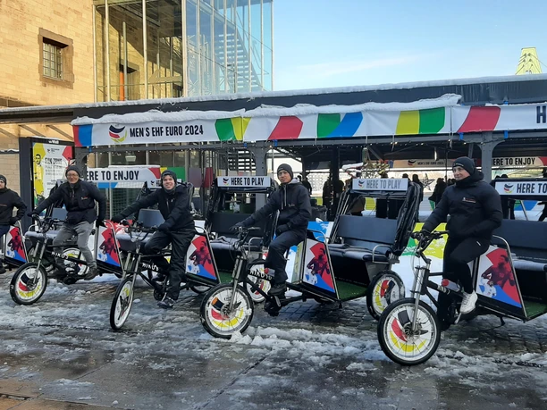 Rikolonia_Event EHL.jpg Four people ride on bicycle rickshaws in cold weather, surrounded by colorful advertising banners.