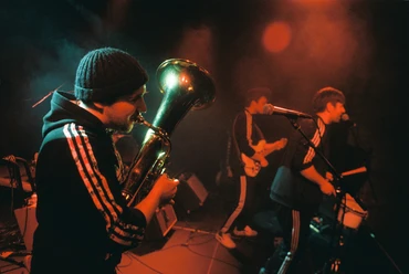 BalkanRockTrio-Pressefoto2-Querformat-Credit-Julian-Wieder.jpg Drei Musiker spielen auf einer Bühne, ein Tuba-Spieler im Vordergrund, beleuchtet von warmem Licht.
