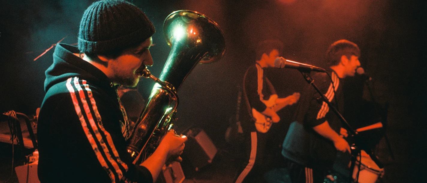 BalkanRockTrio-Pressefoto2-Querformat-Credit-Julian-Wieder.jpg Drei Musiker spielen auf einer Bühne, ein Tuba-Spieler im Vordergrund, beleuchtet von warmem Licht.