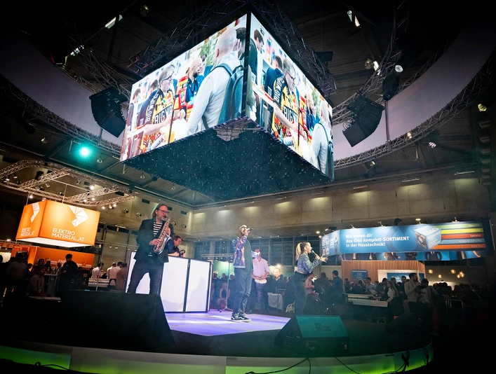 Frauenthal-Expo-Wien-PLUS-Messeband-Entertainment-DJplus-04.jpg Musiker auf einer Messebühne, große Bildschirme zeigen lebhafte Szenen, umgeben von Publikum.</p>Musicians on an exhibition stage, large screens show lively scenes, surrounded by an audience.</p> <p
