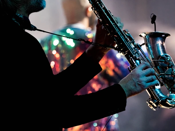 PLUS-Messeband-Entertainment-DJplus-16.jpg Ein Musiker in dunklem Outfit spielt leidenschaftlich Saxophon auf einer stimmungsvollen Bühne.</p>
