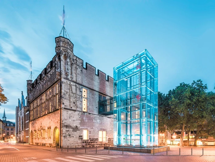 Gürzenich Köln, Außenansicht Altes Rathaus mit modernem gläsernen Bau im Abendlicht, umgeben von Bäumen und beleuchteter Straße.</p>Old town hall with modern glass building in the evening light, surrounded by trees and illuminated street.</p> <p