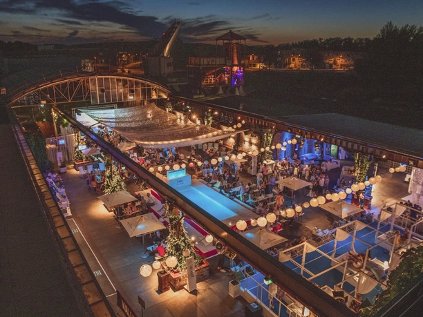 Biergarten der HALLE Tor 2 - Open Air Offene Terrasse eines Restaurants bei Nacht mit stimmungsvoller Beleuchtung und vielen Gästen.