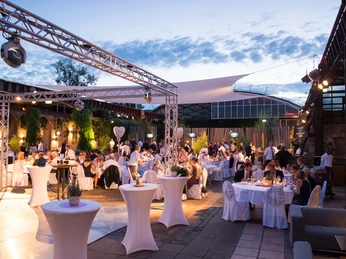 Biergarten der HALLE Tor 2 - Open Air.jpg Festlich gedeckter Außenbereich einer Veranstaltung bei Dämmerung, beleuchtet von warmem Licht.</p>Festively covered outdoor area of an event at dusk, illuminated by warm light.</p> <p