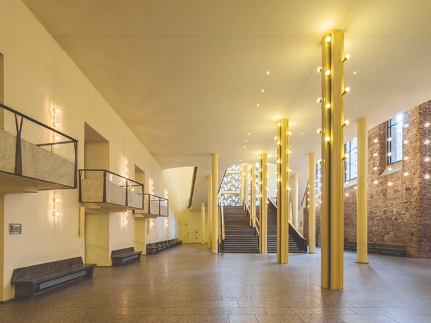 Gürzenich Köln, Erdgeschoss-Foyer Leerer, lichtdurchfluteter Saal mit Marmorelementen, Treppenaufgang und modernen Leuchten.