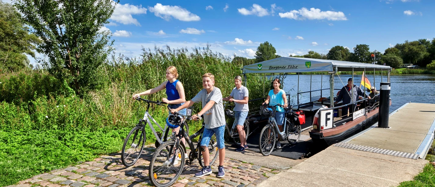 Bargener Fähre Radfahrende steigen an einer kleinen Flussfähre bei sonnigem Wetter ans Ufer.