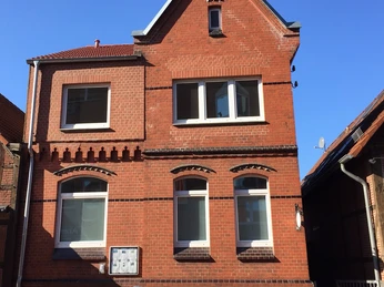 Büro außen.jpg Zweistöckiges Backsteingebäude mit vier Fenstern und einem spitzen Giebel unter blauem Himmel.