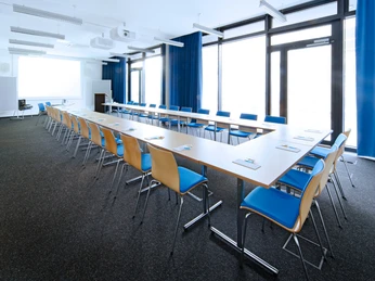 ZMMK-Seminarraum-U-Form Moderner, großzügiger Seminarraum mit U-förmig angeordneten Tischen und blauen Stühlen.</p>Modern, spacious seminar room with U-shaped tables and blue chairs</p>.