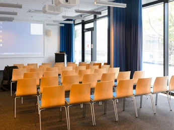 ZMMK-Seminarraum-Theaterbestuhlung Moderner Konferenzraum mit Holzstühlen, blauem Vorhang und großer Leinwand.</p>Modern conference room with wooden chairs, blue curtain and large screen.</p> <p
