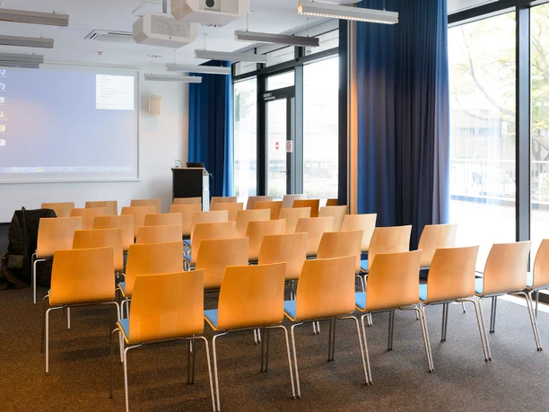 ZMMK-Seminarraum-Theaterbestuhlung Moderner Konferenzraum mit Holzstühlen, blauem Vorhang und großer Leinwand.</p>