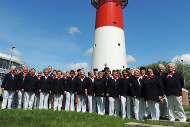 Nordseechor in Büsum Eine Gruppe Menschen in weißen Hosen und blauen Oberteilen. Im Hintergrund ein Leuchtturm.