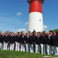 Nordseechor in Büsum Eine Gruppe Menschen in weißen Hosen und blauen Oberteilen. Im Hintergrund ein Leuchtturm.