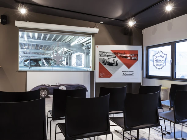 _MG_0461.jpg Das Bild zeigt einen Raum mit schwarzen Stühlen, einem Fenster und einer Wanddekoration mit Auto-Motiven.