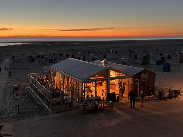 The picture shows the 'Sonnendeck Borkum' on the beach under a colorful sunset. People enjoy the cozy atmosphere of the illuminated pavilion with a view of the sea. The appealing ambiance invites you to linger.