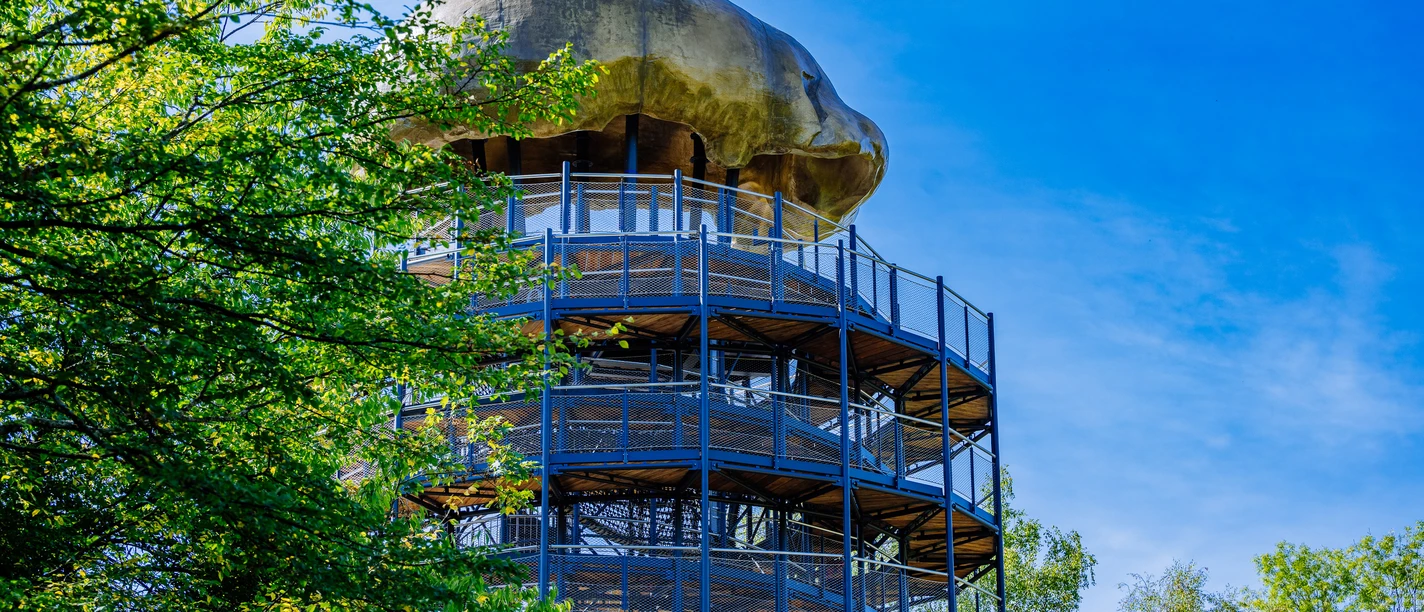 Führung Fundstelle & Erlebnisturm Höhlenblick - Neanderthal Museum Rundturm aus Metall und Holz in bewaldetem Gebiet mit auffälligem pilzförmigem Dach gegen blauen Himmel.