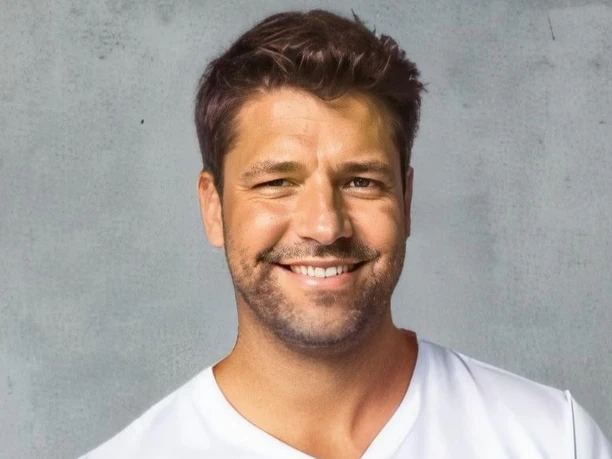 Foto.jpg Ein lächelnder Mann mit braunem Haar im weißen T-Shirt vor einer grauen, strukturierten Wand.