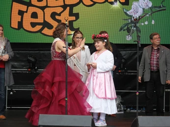 Zwei Mädchen in Festkleidern auf einer Bühne bei einem Obstfest, begleitet von Erwachsenen.