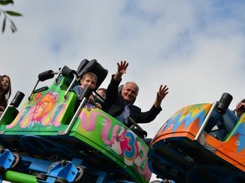 Ein älterer Herr und ein Junge fahren lächelnd auf einer bunten Achterbahn, umgeben von blauem Himmel.