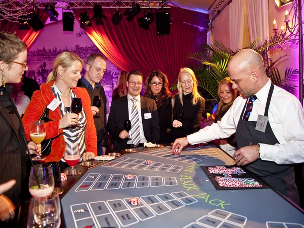 Casino Vinophil. Wein Black-Jack Tisch People in evening dress are standing at an elegant blackjack table in a stylish casino, enjoying wine. A professional croupier deals the cards with a concentrated gaze.