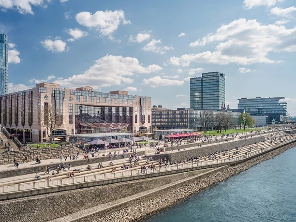 Außenansicht Uferpromenade am Rhein an einem sonnigen Tag, belebt mit Menschen, moderner Architektur im Hintergrund.</p>