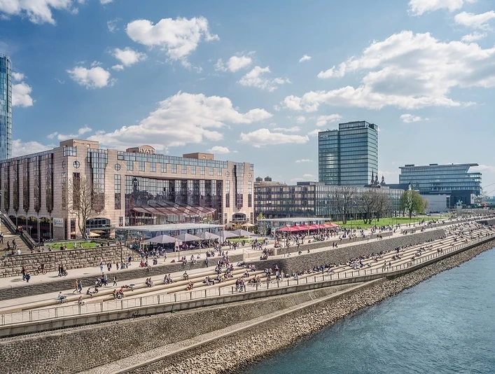 Außenansicht Uferpromenade am Rhein an einem sonnigen Tag, belebt mit Menschen, moderner Architektur im Hintergrund.</p>Riverside promenade on the Rhine on a sunny day, bustling with people, modern architecture in the background.</p> <p