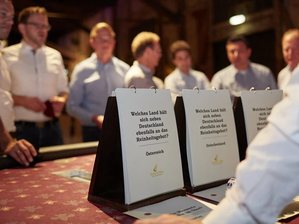 Hopfen & Malz. Genuss-Quiz Tisch <p>Personen diskutieren an einem Tisch über Quizfragen in einem stimmungsvoll beleuchteten Raum.</p>