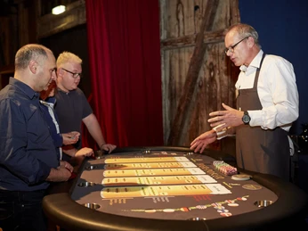 Hopfen & Malz. Bier-Poker Tisch Ein Croupier erklärt drei Besuchern das Spiel an einem Casino-Tisch in einem stimmungsvollen Raum.</p>A croupier explains the game to three visitors at a casino table in an atmospheric room.</p> <p