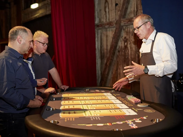 Hopfen & Malz. Bier-Poker Tisch A croupier explains the game to three visitors at a casino table in an atmospheric room.</p> <p